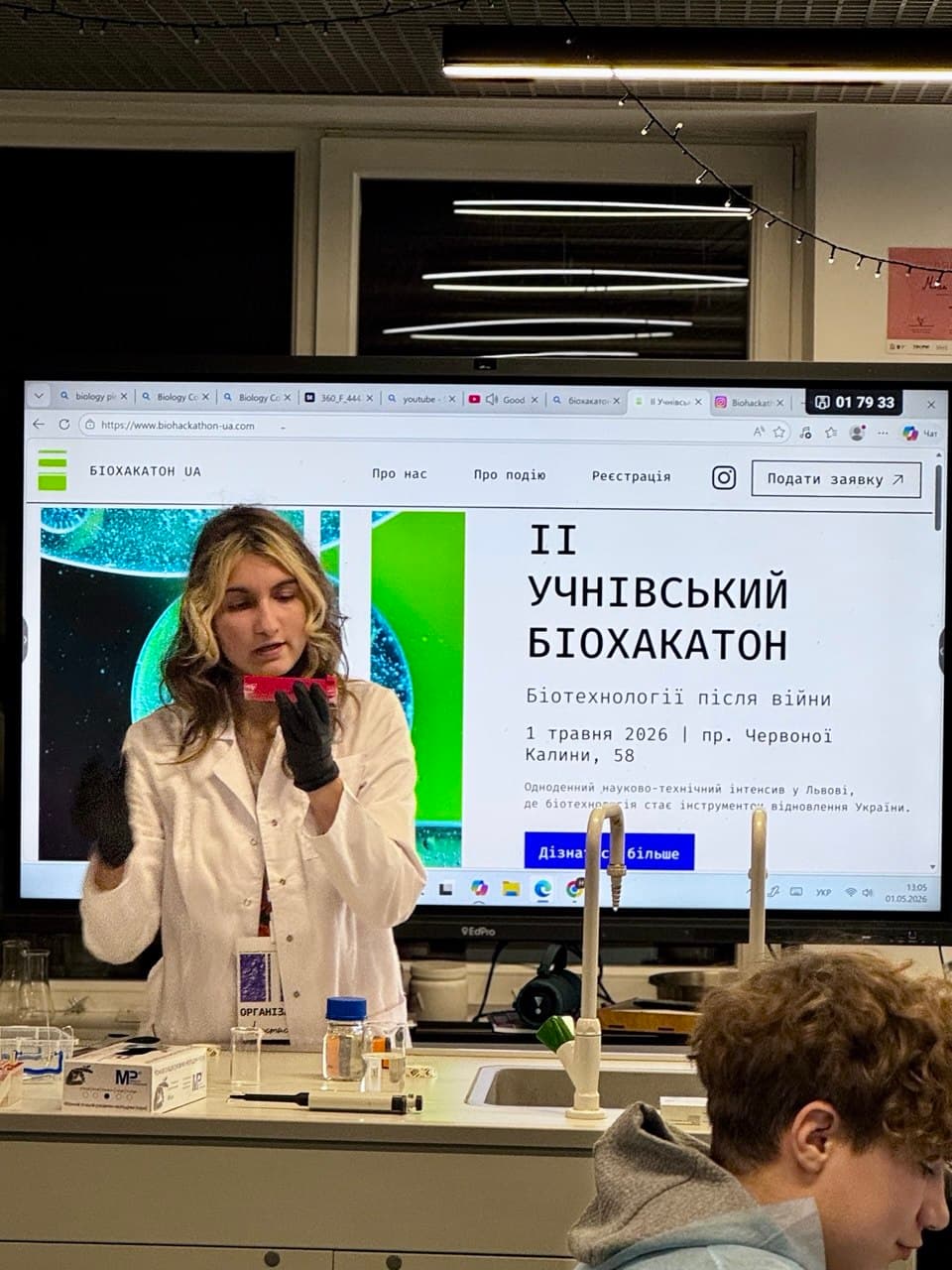 Доповідачка в лабораторному халаті під час демонстрації на тлі презентації біохакатону